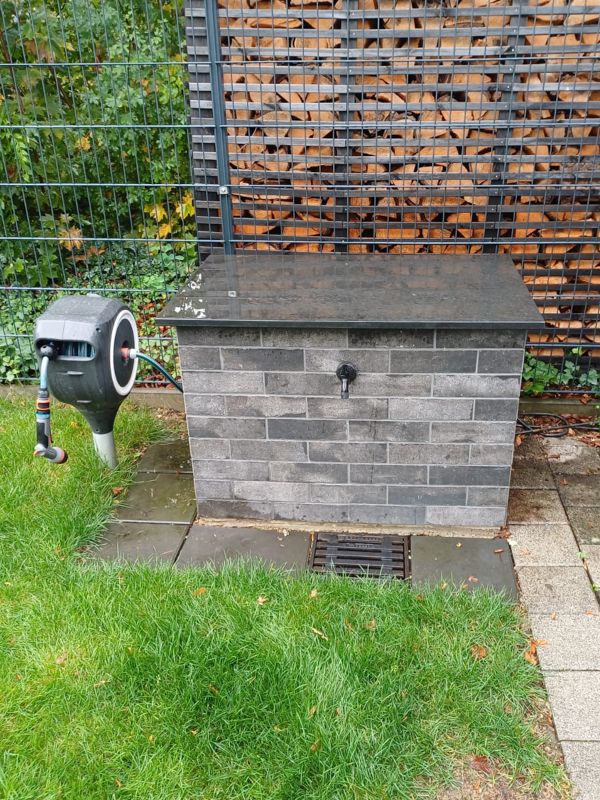 Gartenpumpengehäuse mit grauer Verkleidung in Steinoptik, dunkler Abdeckung und Wasserhahn