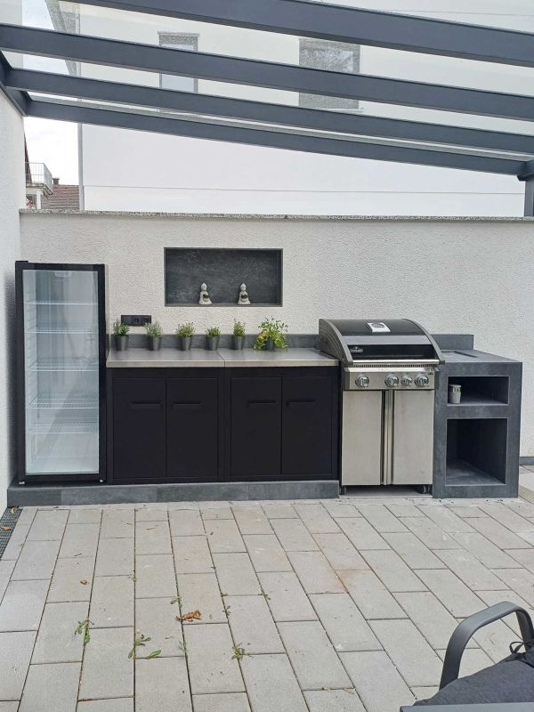 Überdachte Outdoorküche mit Grill, Spüle, dunklen Unterschränken und verputzter Rückwand