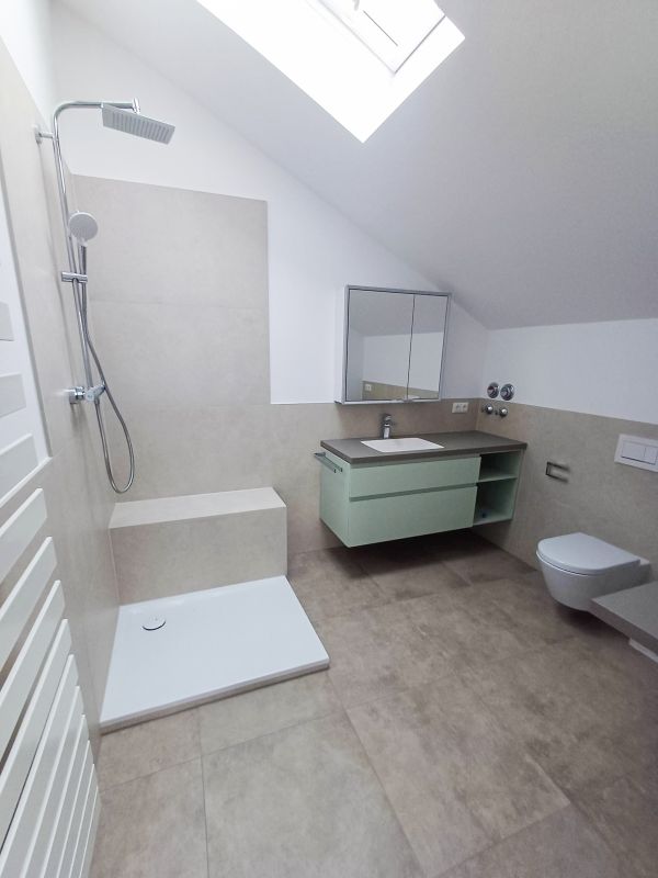Badezimmer unter einer Dachschräge mit Dusche, grauen Bodenfliesen, grünem Waschtischunterschrank und wandhängender Toilette