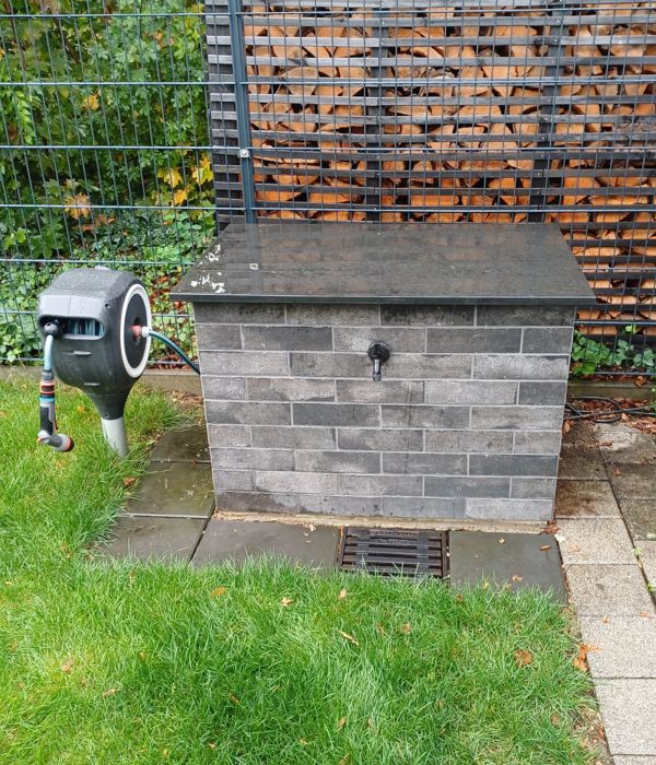 Gartenpumpengehäuse mit grauer Verkleidung in Steinoptik, dunkler Abdeckung und Wasserhahn