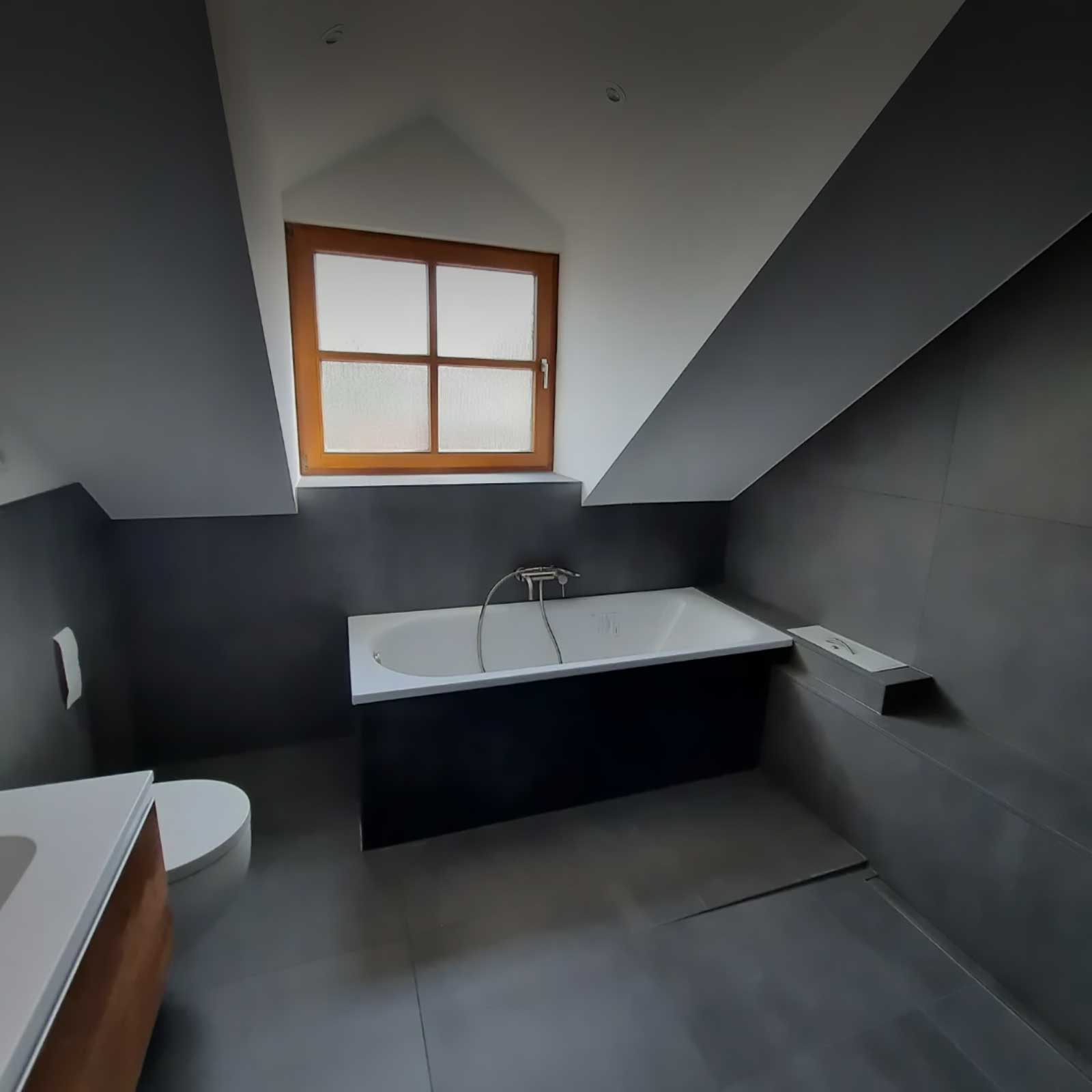 Badezimmer unter einer Dachschräge mit Badewanne unter dem Fenster und großformatigen grauen Wand- und Bodenfliesen